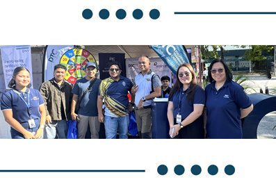 Group of smiling people at an outdoor event, many wearing blue shirts with lanyards, posing in front of a dartboard and banners.