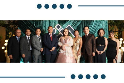 Ten formally dressed individuals posing together for a group photo at a gala, standing in front of a teal decorative backdrop with lights and geometric decor.