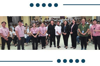 Group of people in an office hallway posing for a team photo; several in pink shirts, others in black or business attire.