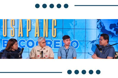 Panel discussion with four speakers seated in a row on a stage, each with a microphone, against a blue map-themed backdrop.