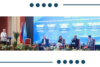 Panel discussion at a conference: five experts seated on stage with a podium speaker and flags behind them on a bright blue backdrop.