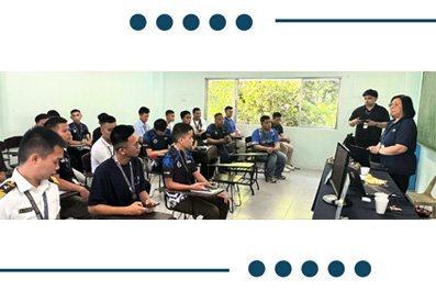 Teacher explains material at the front of a classroom as students sit at desks facing a computer monitor.