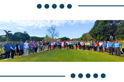 A large group of people standing in a line on a grassy field outdoors, posing for a group photo on a sunny day.