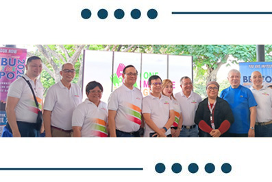 Group of people posing for a group photo at an outdoor community event, some wearing white shirts with colorful stripes, with banners in the background.