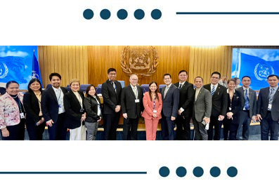 Group of about 15 professionals in business attire posing for a photo in a formal setting with a wooden backdrop and flags in the background.