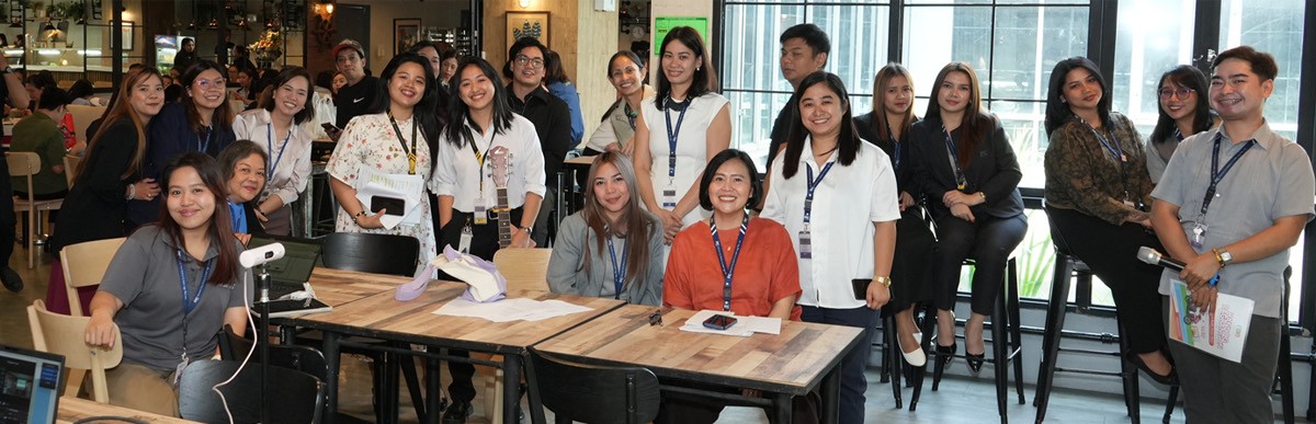 Group of coworkers posing for a photo around a long wooden table in a modern office cafe, many wearing lanyards and smiling at the camera.