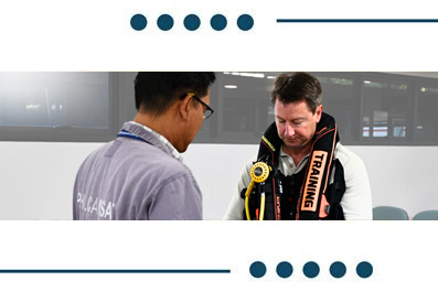 Two men in a training session; one wears a safety harness labeled TRAINING and talks with the other in a gray shirt in a modern workspace.