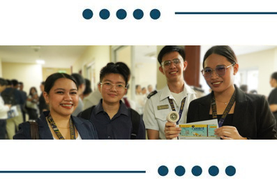Four young adults pose and smile for a group photo at an event in a hallway, with one person showing a colorful card.