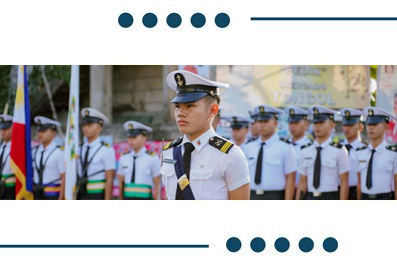 Young military officer in a white uniform leads a line of cadets in formation during an outdoor parade, with flags and bystanders in the background.