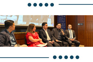 Panel discussion with five people seated on a stage, including a woman in red among men in suits; backdrop features wood paneling and a projector screen.