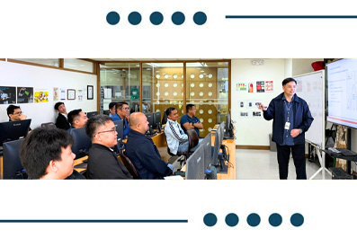 Group of professionals watching a presenter give a talk at a whiteboard in a modern classroom/office setting.