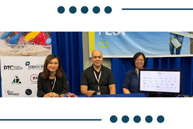 Three people sit at a blue trade‑show booth with a display monitor on the right and sponsor logos on a backdrop.