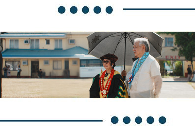 Two people in ceremonial outfits walk outdoors under a large umbrella, with a light-blue building in the background.