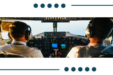 Two pilots wearing headsets in the cockpit, seen from behind, with the instrument panel and controls ahead.