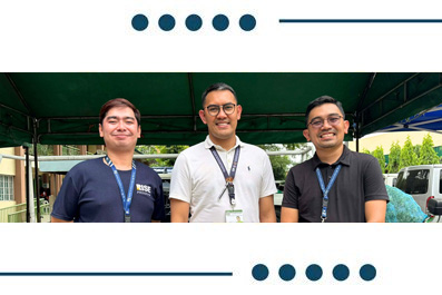 Three men with lanyards standing side by side under a green canopy at an outdoor event.