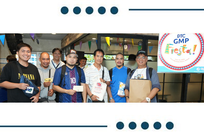 Group of people posing with bags and papers at an indoor GMP Fiesta event, banner overhead and logo on the right.