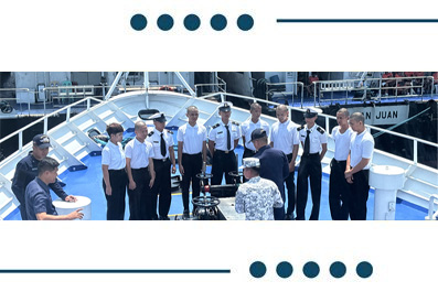 Group of uniformed ship crew standing in a semicircle on a boat deck, listening to an officer addressing them, with harbor background.