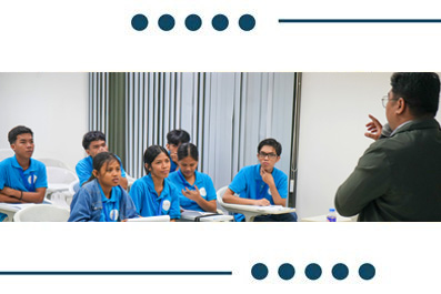 Instructor giving a lecture to a class of students in blue uniforms seated at desks in a bright classroom.