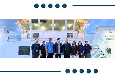 Eight people stand in a row on the deck of a white ship, posing for a group photo; a 'Safety First' sign is visible on the vessel.