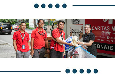 Four volunteers in red shirts with lanyards hand a donation bag to a woman beside a Caritas charity vehicle.